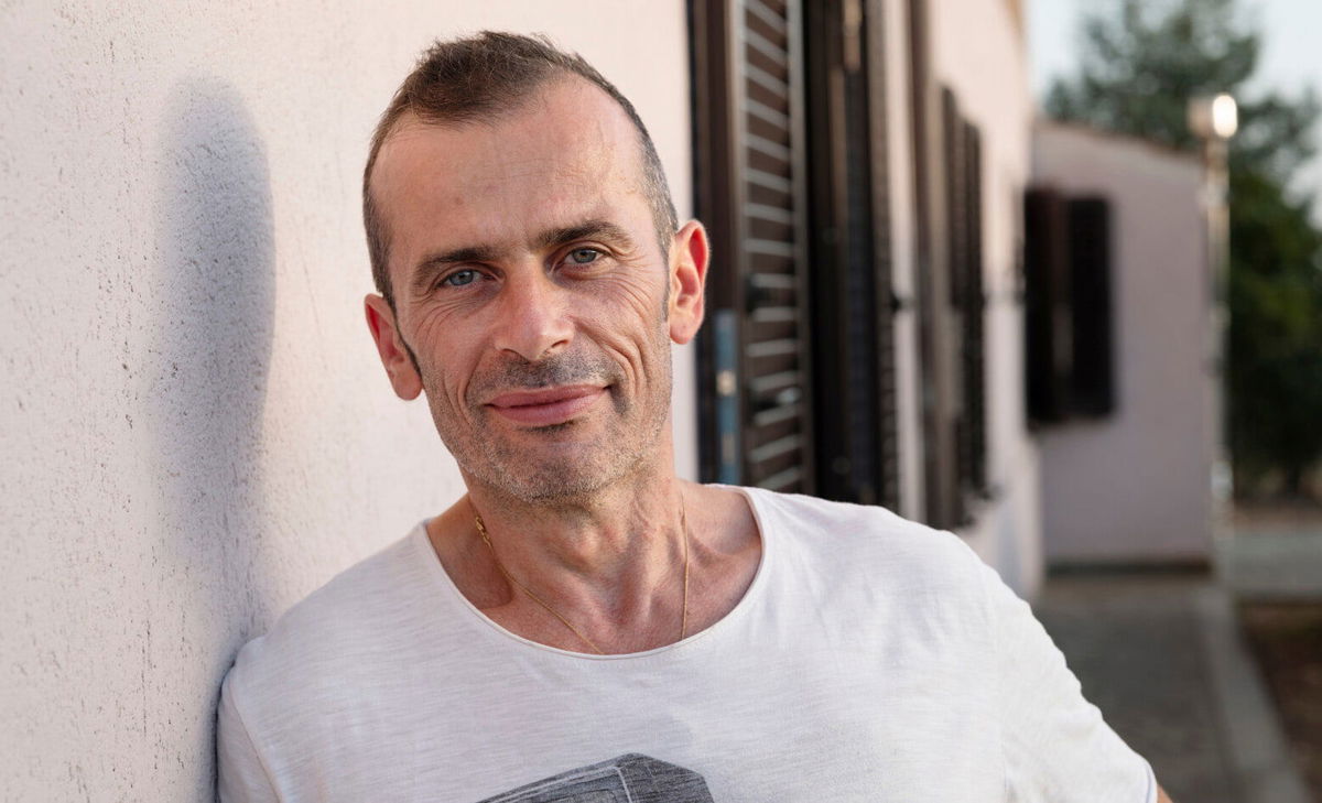 Portrait photo of Andrea stood outside his house. Andrea lives with MS and wishes he had known a little more about what to expect with the associated bladder and bowel issues.  
