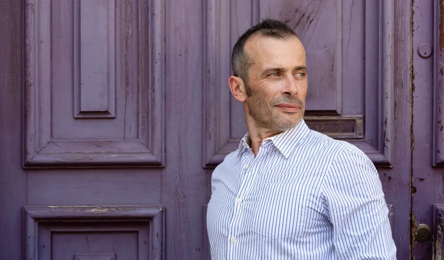 Portrait photo of Andrea stood in front of a purple door. Andrea was diagnosed with progressive remitting multiple sclerosis and experienced problems with both his bladder and bowel. 