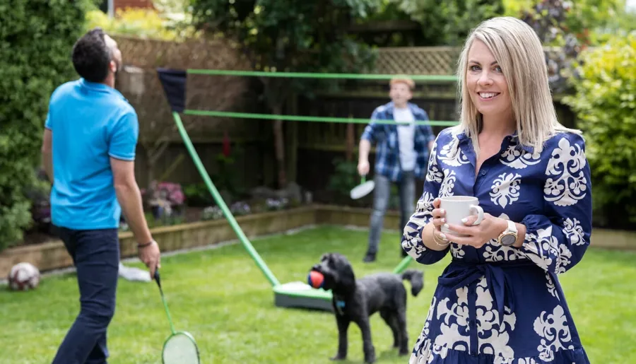 Laura stands in her garden with her family playing badminton in the background. Laura uses Peristeen Plus to help manage her bowel issues which result from her MS.  